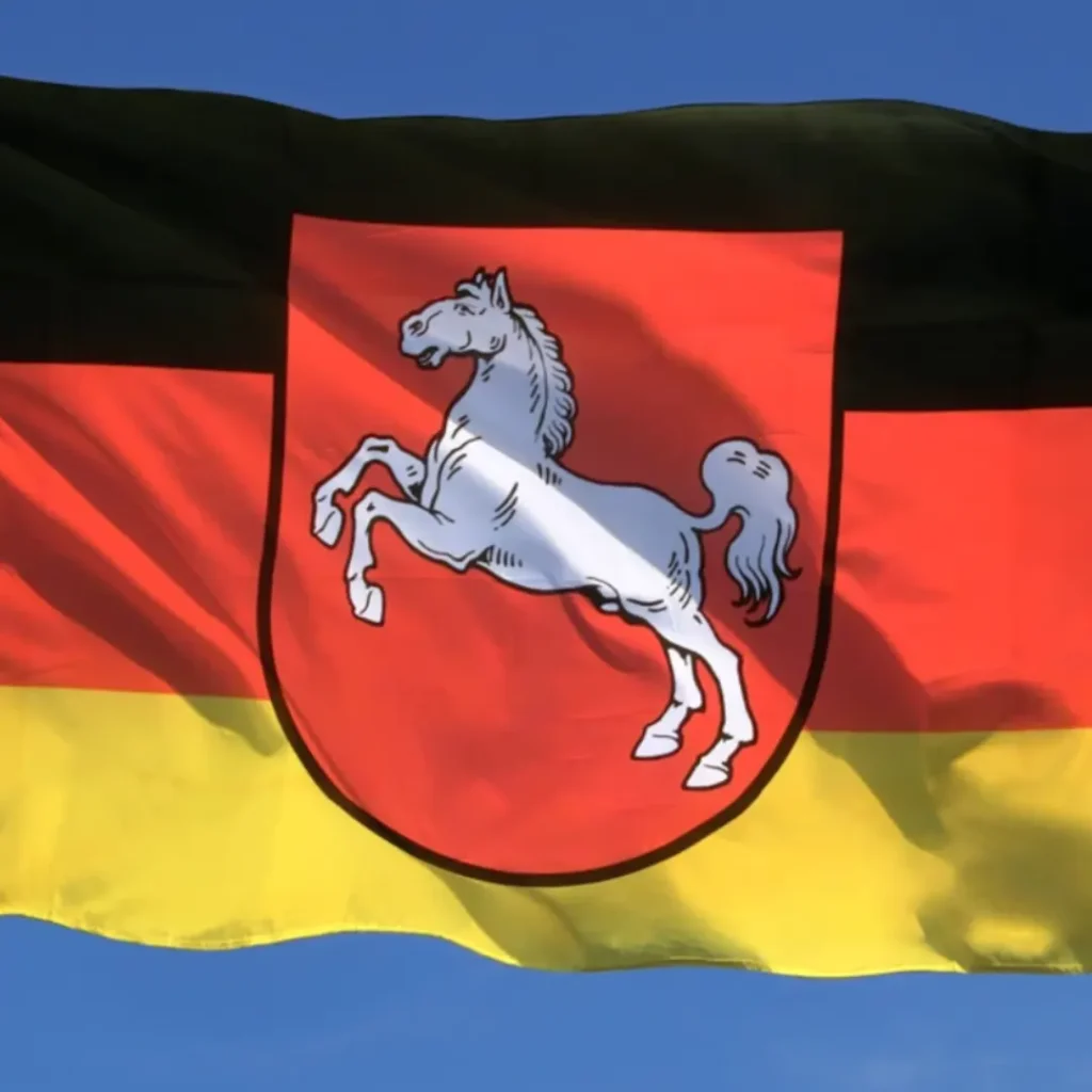 Das Bild zeigt die Fahne des Bundeslandes Niedersachsen. Im Hintergrund ist wolkenloser Himmel sichtbar.