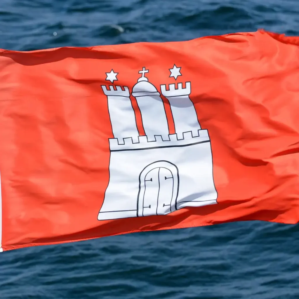 Das Bild zeigt die wehende Fahne der Freien und Hansestadt Hamburg. Im Hintergrund ist Wasser sichtbar.