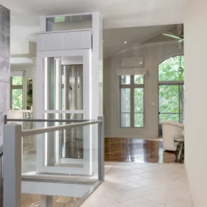 Das Bild zeigt die obere Haltestelle eines Luma-Senkrechtliftes inkl. Fahrstuhlkabine voneinem Standort aus, der hinter dem Fahrstuhl liegt. Sehr gut erkennbar sind die Glaselemente und die lichtdurchflutete Kabine.